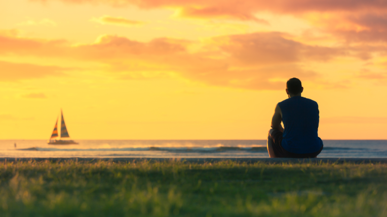 Man at a lake, sitting on a cliff watching the sun rise with orange sky.